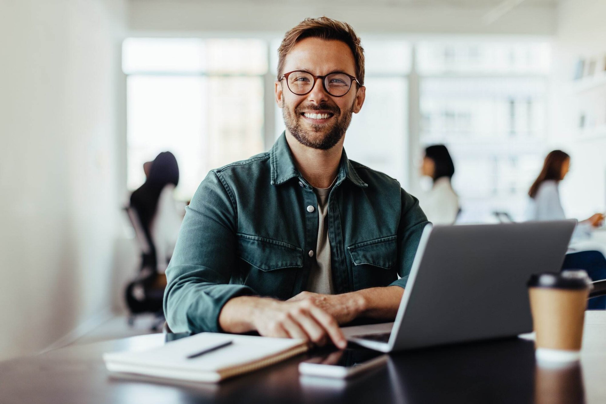 Smilende mand med briller sidder ved skrivebord på kontoret og bruger tidsregistrering en computer.